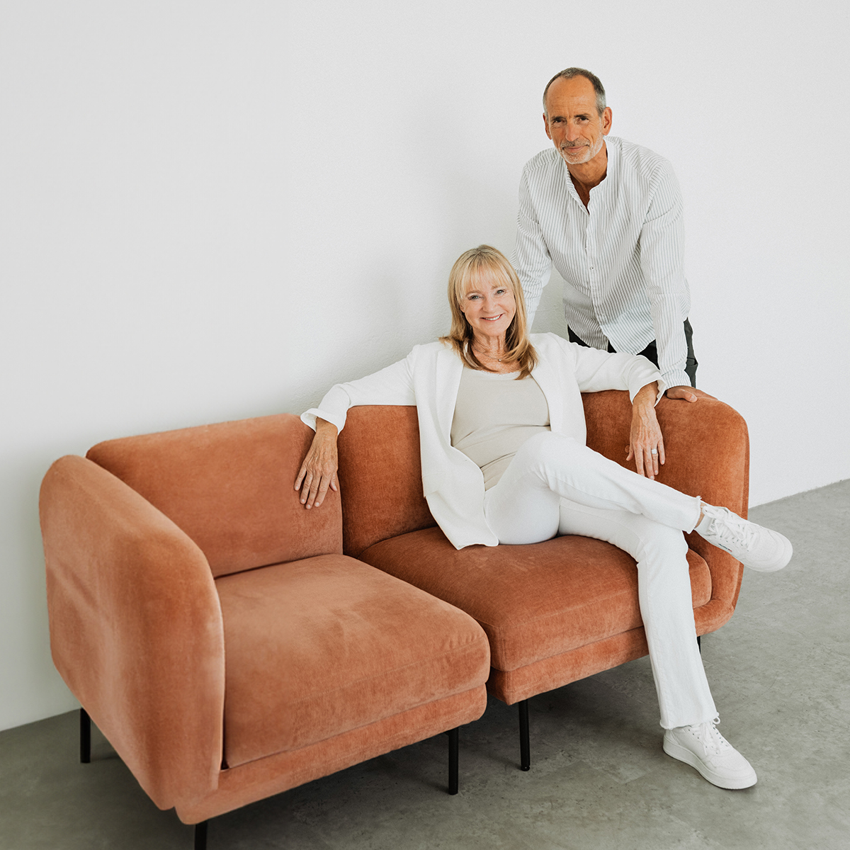 Auf einem terracottafarbenen Sofa sitzt Dr. med. Petra Bracht. Ihr Mann Roland Liebscher-Bracht steht hinter ihr und dem Sofa.