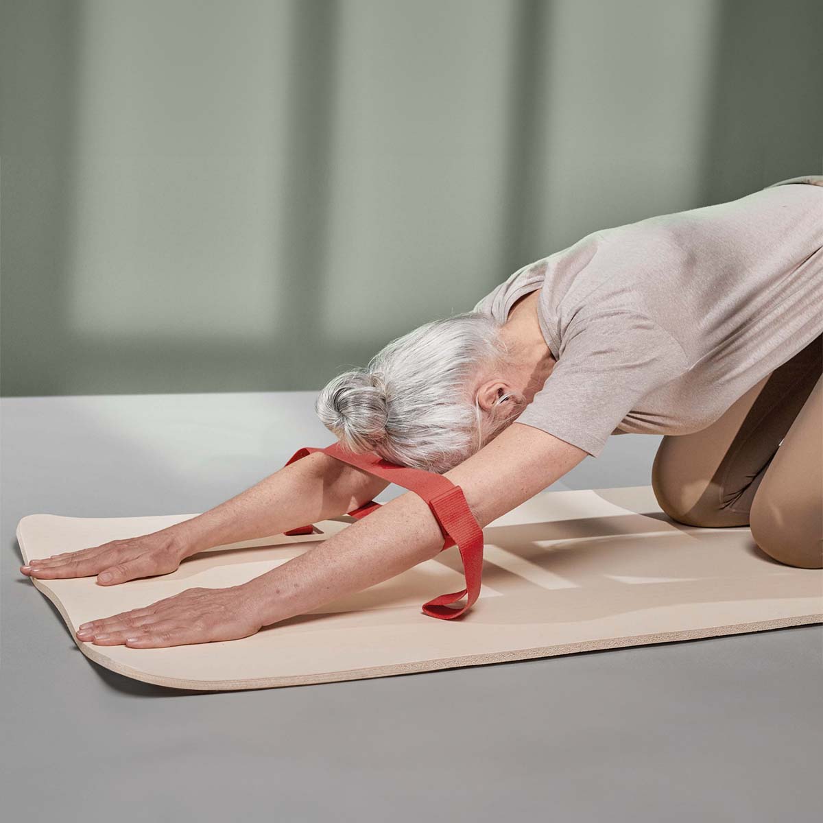 Eine Frau kniet auf einer Übungsmatte. Die Arme  liegen am Boden ab. Die rote Übungsschlaufe von Liebscher & Bracht ist um die Oberarme gewickelt, um die gewünschte Dehnung  im Schulterbereich zu unterstützen.